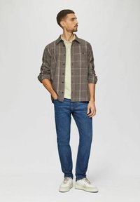 Grau kariertes Hemd über einem grünen T-Shirt, kombiniert mit blauen Jeans und weißen Sneakern mit beigen Akzenten. Das Model steht mit einer Hand in der Tasche.