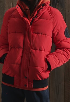 Osoba ubrana w czerwoną pikowaną kurtkę z czarnymi mankietami i obszyciem, z rękami w kieszeniach, stojąca na tle drewnianej ściany.