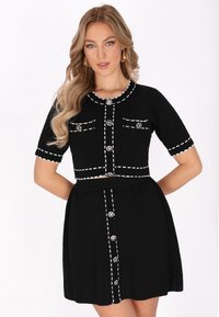 Schwarzes Kleid mit einem figurbetonten strukturierten Oberteil, kurzen Ärmeln und einem ausgestellten Rock. Mit weißen Steppnähten und dekorativen Knöpfen erhältlich.