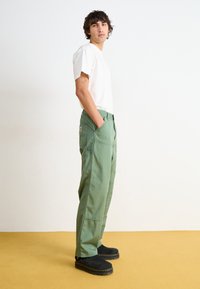 Pantalons cargo verts avec poches latérales, coupe décontractée et étiquette sur la taille. Associés à un t-shirt blanc à manches courtes et des sabots noirs.