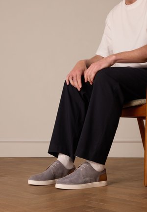 Grijze suède sneakers met bruine leren accenten, witte veters en een gestructureerd oppervlak. Draagt witte sokken en donkere broek.