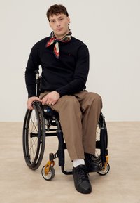 Homme assis en fauteuil roulant portant une chemise noire à manches longues, un pantalon marron, des chaussures noires, des chaussettes blanches et une écharpe multicolore autour du cou.