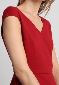 Punainen mekko, jossa on V-pääntie, lyhyet hihat ja istuva vartalo-osa, valmistettu sileästä ja teksturoidusta kankaasta. Siistit linjat korostavat muotoiltua siluettia.
