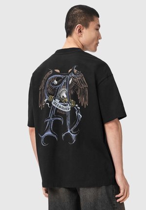 Homme portant un t-shirt noir avec un grand "A" stylisé et un aigle tenant une bannière avec "ALLSAINTS" dans le dos, vu de profil.