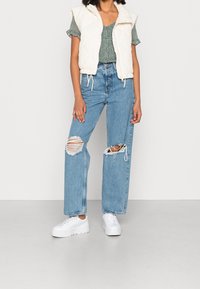 Gilet bianco trapuntato con cappuccio, top verde a pois, jeans blu svasati e strappati, e sneakers bianche. Tessuto texturizzato e strappi visibili.