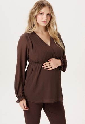Femme aux longs cheveux blonds portant un chemisier marron à col en V et un pantalon, tenant son ventre d'une main.