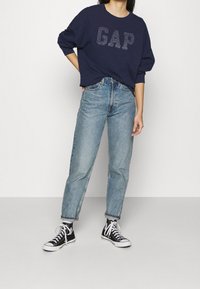 Tmavě modrá mikina s logem "GAP", doplněná světle modrými rovnými džínami a černými vysokými teniskami s bílými doplňky.