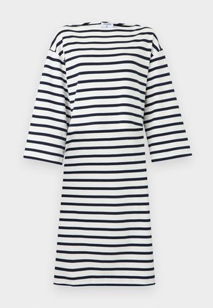 Kvinde stående med hånd på hoften iført knælængde, hvid kjole med lange ærmer med vandrette sorte striber og sorte flade sko.