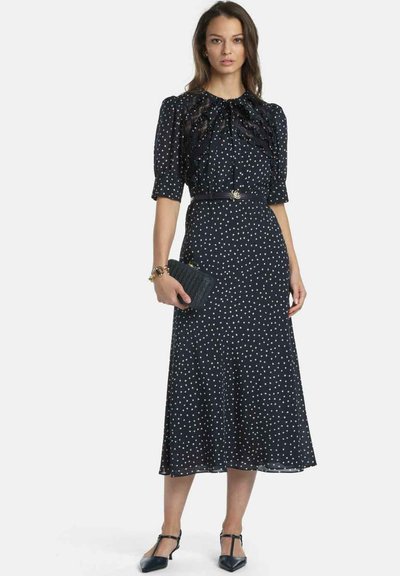 Marineblaues Kleid mit weißen Polka-Dots, kurzen Puffärmeln und einem gerafften Ausschnitt. Es verfügt über eine taillierte Passform mit einem dünnen Gürtel und einem fließenden Rock.