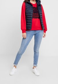 Rode sweatshirt met zwart paillettenlogo, marineblauwe gewatteerde vest, lichtblauwe skinny jeans en witte sneakers. Casual gelaagde outfit.