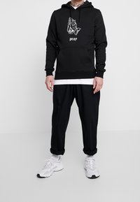 Muže ve černé mikině s kapucí, na které jsou zobrazeny modlící se ruce a text "pray", černé kalhoty s manžetami, bílá vrstvená košile, bílé ponožky a bílé tenisky.