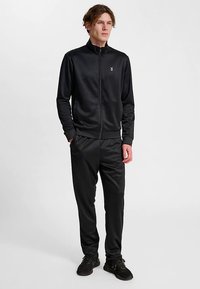 Czarny sportowy dres z kurtką na zamek i dopasowanymi spodniami. Charakteryzuje się gładką fakturą, dopasowanym krojem oraz małym emblematem na kurtce.