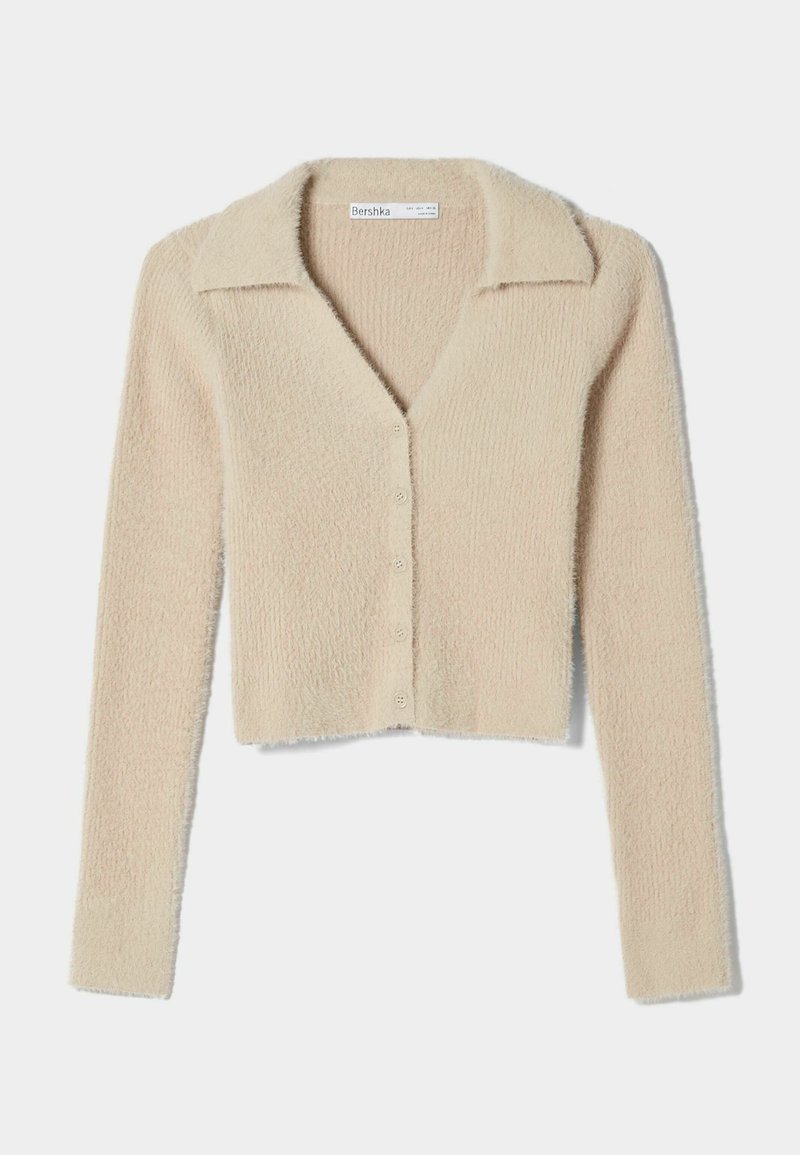 Beige kort cardigan tillverkad av mjukt, texturerat tyg, med knäppning framtill, v-ringning och långa ärmar med klassisk krage.