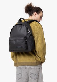 Mochila negra hecha de un material suave, con un bolsillo frontal con cremallera, correas ajustables y un parche de marca, llevada sobre un suéter amarillo.