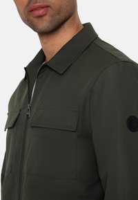 Veste vert foncé en tissu lisse, avec une fermeture éclair, deux poches poitrine et un emblème rond noir sur la manche.