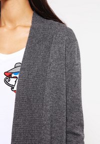 Cardigan gris tricoté avec texture côtelée, design à ouverture sur le devant et manches longues, superposé sur une chemise blanche avec un motif graphique coloré.