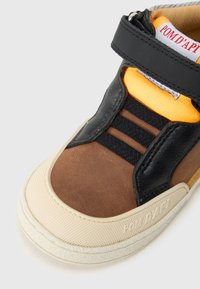 Ténis de criança com painéis castanhos, pretos e laranjas, bico texturizado em bege, tira de Velcro e marca "POM D’API" na sola e na língua.
