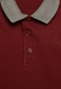 Polo shirt bordeaux con colletto grigio lavorato a maglia e tre bottoni marroni. Presenta una texture liscia e un logo ricamato sottile.