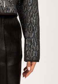 Zwarte cropped jacket met een metallic schubbenpatroon in tinten blauw en zilver. De mouwov heeft een effen zwarte rand. Gecombineerd met een zwarte leren rok.