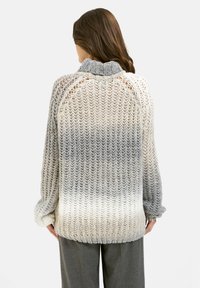 Frau, die mit dem Rücken zur Kamera steht, trägt einen dick gestrickten Ombre-Pullover in Grautönen und Creme mit geripptem Kragen sowie grauen Hosen.