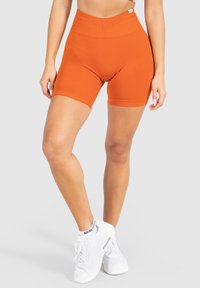 Shorts de vélo taille haute orange en tissu extensible, avec une texture lisse et une coupe ajustée, associés à des baskets blanches.