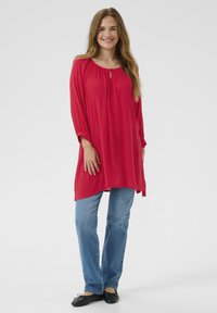 Haut tunique rouge avec un col rond et un détail en forme de clé, associé à un jean bleu. Le tissu semble doux et léger. Chaussures plates noires.