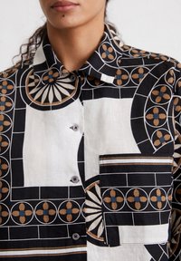Camicia a maniche corte in bianco e nero con motivo geometrico, caratterizzata da disegni floreali in marrone e verde teal, e una tasca sul petto con dettagli a righe.