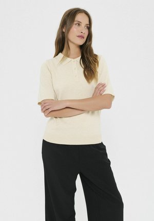 Kvinde med langt brunt hår iført en beige kortærmet polo-skjorte og sorte bukser, stående med armene krydset mod en neutral baggrund.
