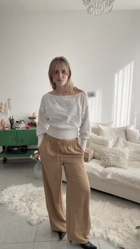 Νεαρή γυναίκα με μακριά ξανθά μαλλιά, φορώντας λευκό τοπ με μακριά μανίκια και μπεζ παντελόνι φαρδιάς γραμμής, στέκεται σε ένα φωτεινό σαλόνι με λευκό καναπέ και πράσινη ντουλάπα.