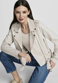 Jasno beżowa skórzana kurtka motocyklowa z srebrnymi detalami, zapinanymi kieszeniami i paskiem w talii. Nosisz ją na beżowej bluzce i dżinsach.