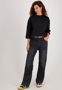 Vrouw in een zwarte langemouwen top, donkere wijde jeans met een patroonriem, en zwart-witte sneakers, staand tegen een effen achtergrond.