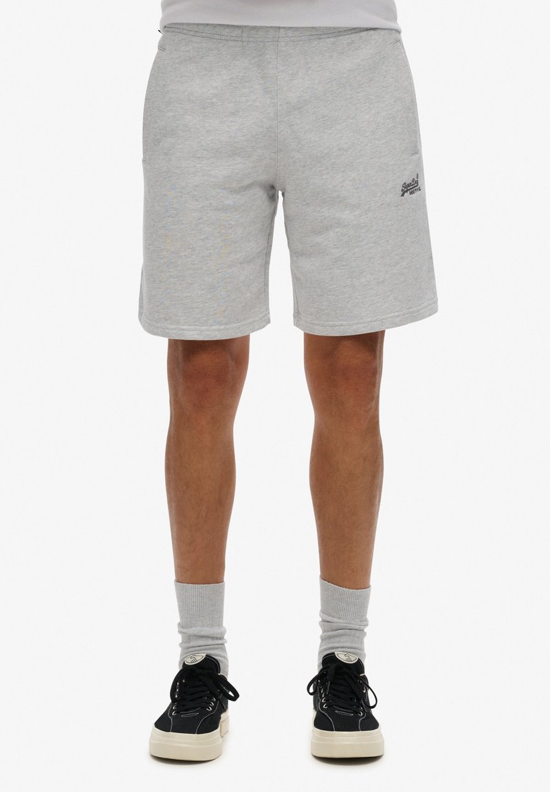 Pantalones cortos de algodón grises con una cintura elástica, bolsillos laterales y un logo impreso. Se llevan con calcetines grises y zapatillas negras.