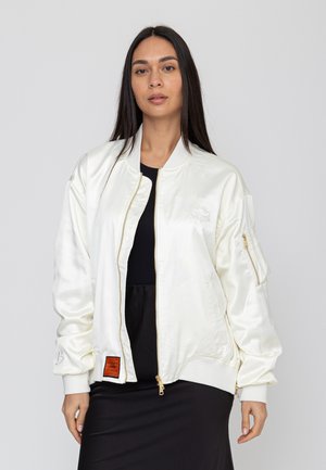 Frau mit langen dunklen Haaren, die eine weiße Satin-Bomberjacke über einem schwarzen Oberteil und Rock trägt, stehend vor einem einfarbigen hellen Hintergrund.