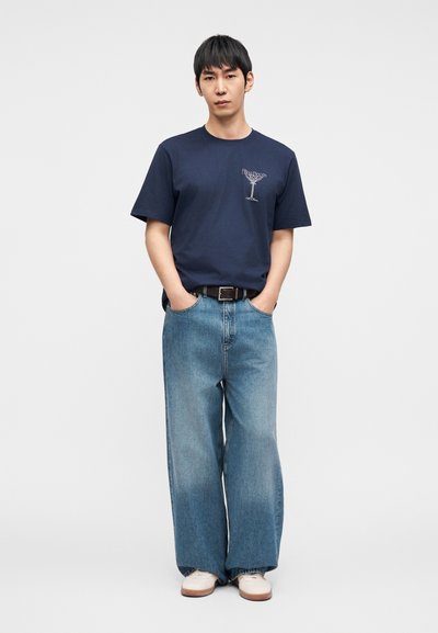 Μπλε ναυτικό βαμβακερό μπλουζάκι με εικόνα ποτηριού κοκτέιλ, συνδυασμένο με ανοιχτό μπλε τζιν παντελόνι φαρδιάς γραμμής. Άσπρα αθλητικά παπούτσια ολοκληρώνουν το ντύσιμο.