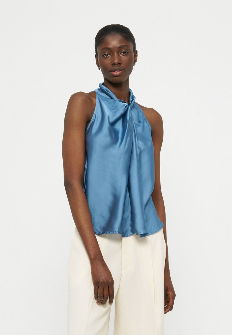 Femme portant un haut bleu en satin à bretelles nouées derrière le cou et un pantalon blanc taille haute, debout devant un fond blanc uni.