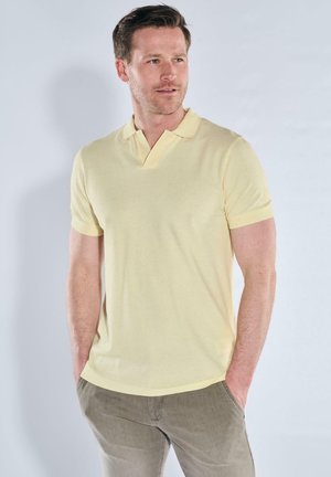 Mann trägt ein hellgelbes, kurzärmliges Poloshirt und graue Hose, steht mit den Händen in den Taschen und schaut leicht nach rechts.