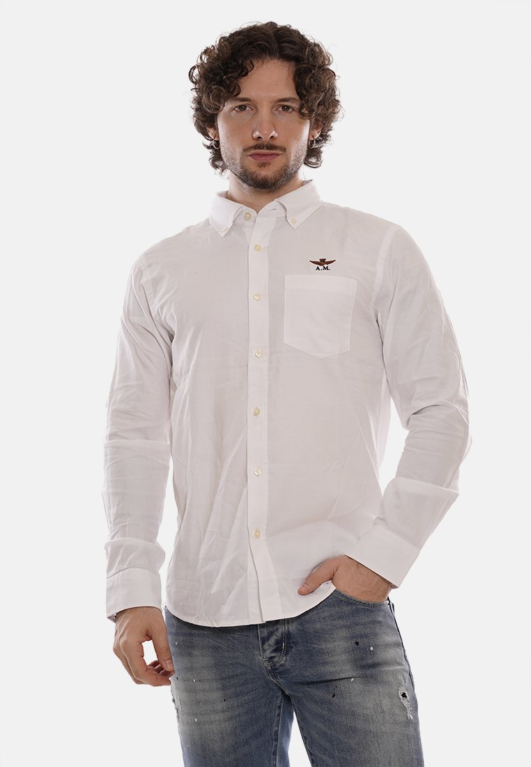 Camicia bianca con bottoni, maniche lunghe, una tasca e un logo ricamato sul petto. Abbinata a jeans blu sbiaditi.