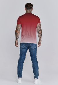 Camiseta de manga corta para hombre en degradado rojo que se desvanece a rosa claro, combinada con jeans azules y zapatillas blancas. Presenta tatuajes visibles en los brazos.