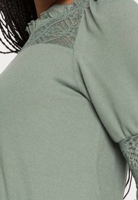 Gros plan d'une personne portant un haut en maille vert sauge avec des détails en dentelle au niveau de l'encolure et des poignets, et des cheveux tressés visibles sur le côté.