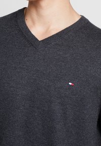 Ciemnoszary sweter w serek wykonany z miękkiego materiału dzianinowego. Posiada małe haftowane logo w kolorach czerwonym, białym i niebieskim na piersi.