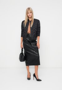 Zwarte jas met zigzagpatroon, tan top, zwarte leren midi rok, en zwarte puntige hakken. Accessoires omvatten een zwarte trekkoordtas.