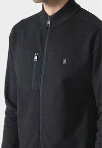 Černá bunda na zip vyrobená z měkké tkaniny, s kapsou na zip na hrudi, vysokým límcem a minimalistickým logem na přední straně.