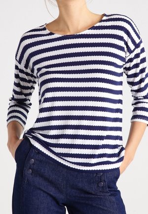 Mujer con camiseta de manga larga a rayas horizontales en azul marino y blanco, metida ligeramente dentro de pantalones azul oscuro con botones.