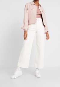 Světle růžová zkrácená bunda, padnoucí žebrovaný top, bílé kalhoty širokého střihu a bílé tenisky vystavené na neutrálním pozadí.