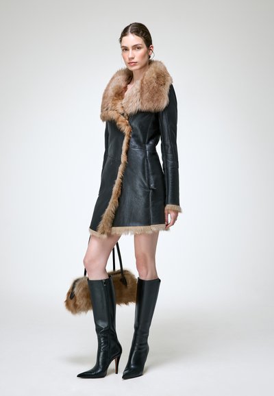 Femme portant un manteau en cuir noir avec bordure en fourrure, des bottes noires à talons hautes jusqu'aux genoux, et tenant un sac à main en fourrure assorti sur un fond uni.
