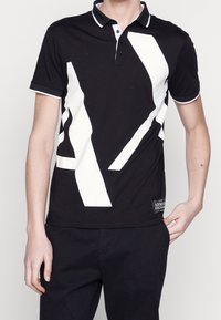 Svart polo t-shirt med djärva vita geometriska mönster, kontrasterande krage och ärmslut, samt en liten märkesetikett vid fållen.