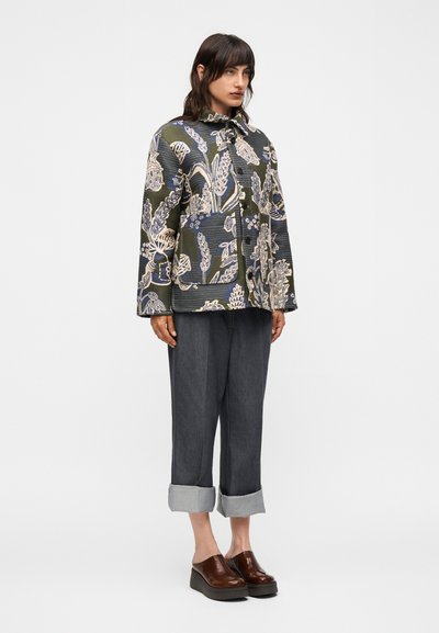 Γυναίκα με μοτίβο σακάκι με κουμπιά, γκρι σκούρο παντελόνι με ρεβέρ και καφέ πλατφόρμες χωρίς κορδόνια, που στέκεται μπροστά σε απλό λευκό φόντο.