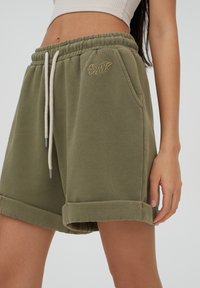 Bermudas de sudadera verde oliva con cinturilla ajustable, dobladillos enrollados, bolsillos laterales y un sutil logo bordado en la parte delantera.