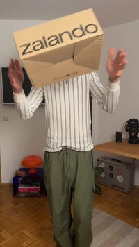 Cutie de carton cu text negru "zalando", ținută de o persoană îmbrăcată cu o cămașă albă cu dungi pe mâneci lungi și pantaloni verzi, stând în interior.