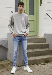 Sweatshirt cinza com texto, jeans justos azuis e ténis brancos. A pé em degraus à frente de uma porta verde, com parede cinza clara.